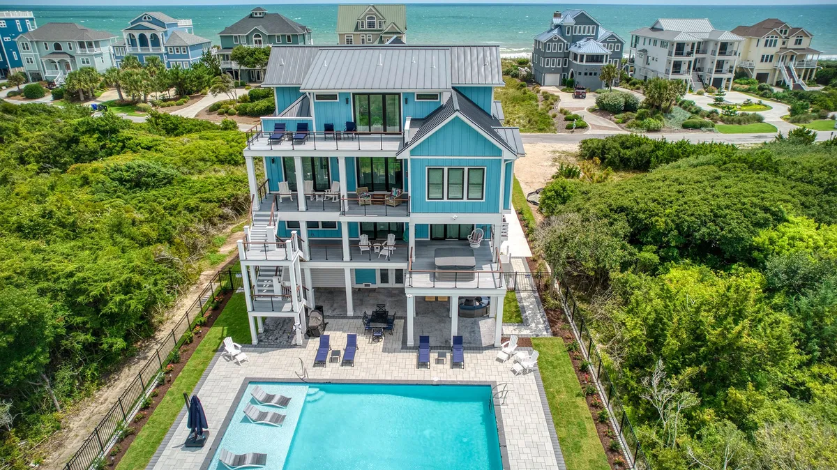 A vibrant exterior scene featuring a sparkling swimming pool surrounded by an outdoor seating area. The colorful house exterior and lush greenery create a lively vacation vibe. The pool area offers views extending toward the ocean, and children can be seen playing nearby. There are also spaces for outdoor dining and relaxing on the balcony.