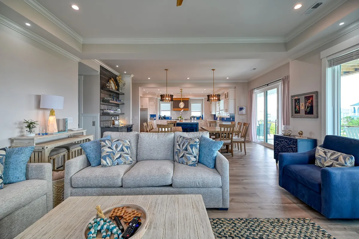 This image showcases a relaxed and inviting open-plan living space featuring a plush sofa with decorative pillows, a prominent dining area, a large kitchen island, and expansive windows that allow natural light to flood the room. The seamless flow between the kitchen, dining, and lounge exemplifies a great room designed for social gatherings, meals, and enjoying the ocean views.