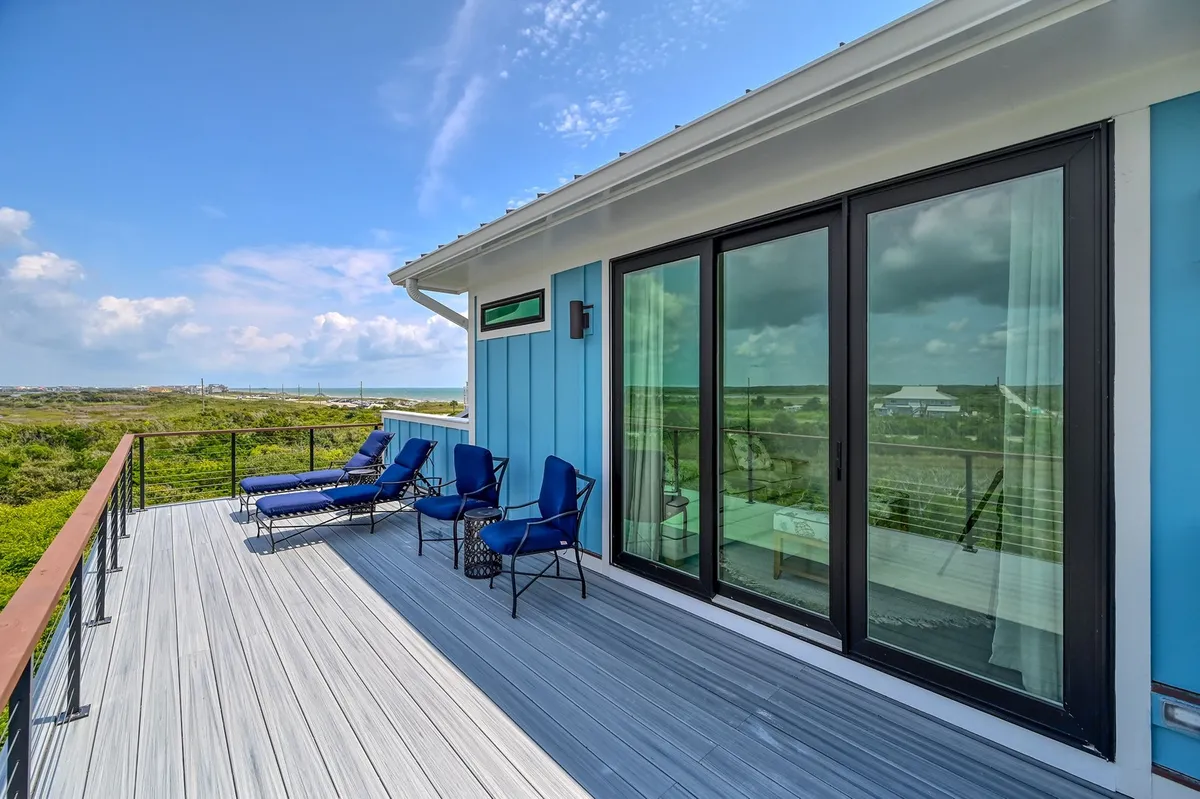 This exterior shot features blue lounge chairs arranged on a patio, facing glass sliding doors that open into the home. In the background, lush green landscaping sets a tranquil, inviting scene. The setup hints at easy transitions between indoor comfort and outdoor relaxation, with plenty of space for group gatherings or quiet moments overlooking the scenery.