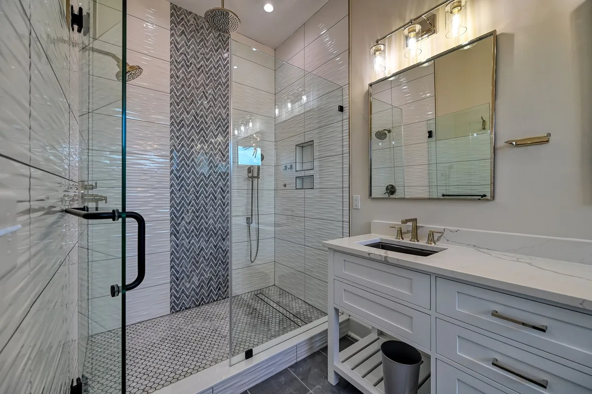 A modern ensuite bathroom with a glass shower enclosure, striking herringbone tile accent wall, and a dual sink vanity. The space exudes a spa-like vibe, perfect for unwinding after a day at the beach.