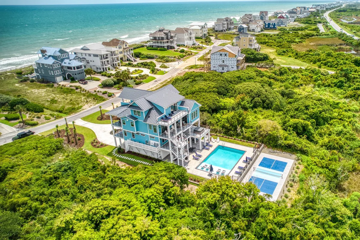 This hero image showcases a stunning, expansive beachfront home with a sparkling private pool, pickleball/tennis courts, and breathtaking views of the ocean at sunset. The home's balconies overlook the pool and the shoreline, inviting guests to imagine relaxing mornings with coffee and fun-filled afternoons in the sun.