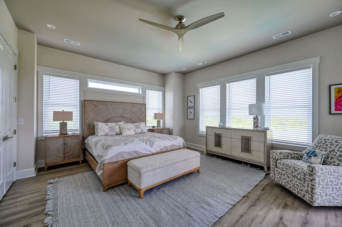 A serene king bedroom featuring a decorative headboard, large windows that flood the space with natural light, a stylish armchair, and a decorative dresser. The ambiance is calm, inviting, and perfectly suited for restful retreats.