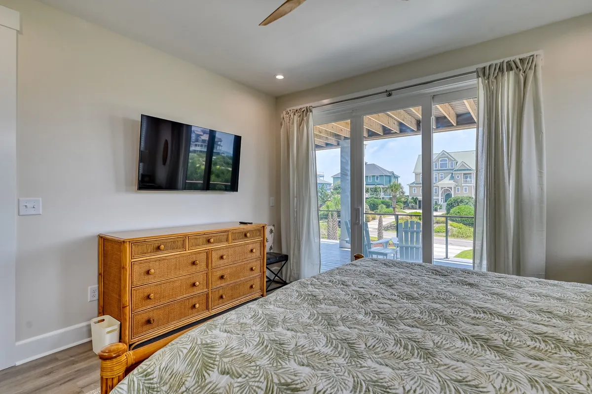 A tranquil king bedroom featuring leafy patterned bedding, a television, dresser, and sliding glass doors that open to a private deck. The space is peaceful, bright, and seamlessly connects to an outdoor retreat.