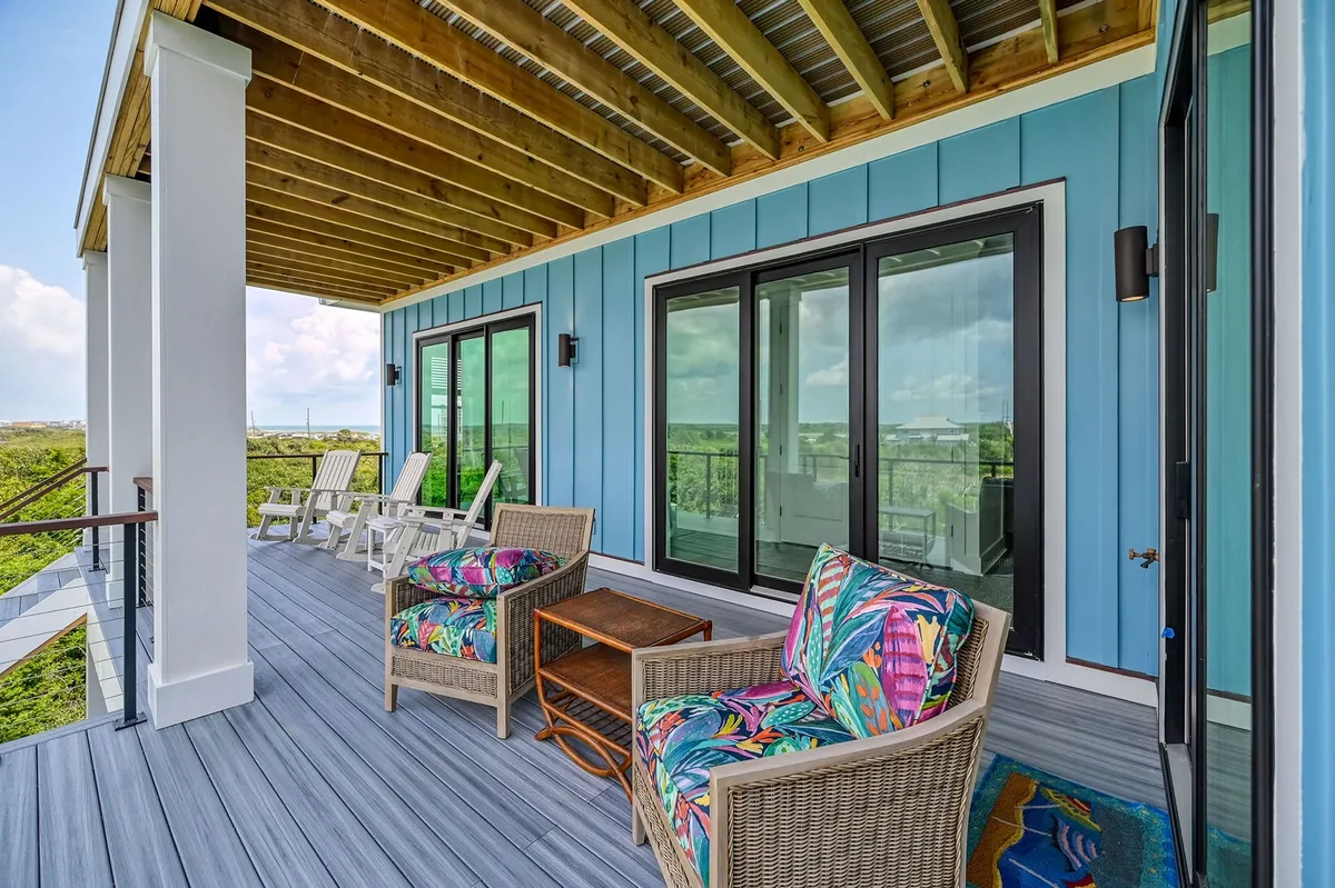 A vibrant exterior dining area featuring colorful chairs arranged around a large wooden table, set against a charming blue wooden wall. The scene exudes a laid-back, welcoming atmosphere perfect for group gatherings or casual meals. The area is ideal for enjoying coffee, reading, or simply relaxing while watching the sunrise or enjoying evening breezes.