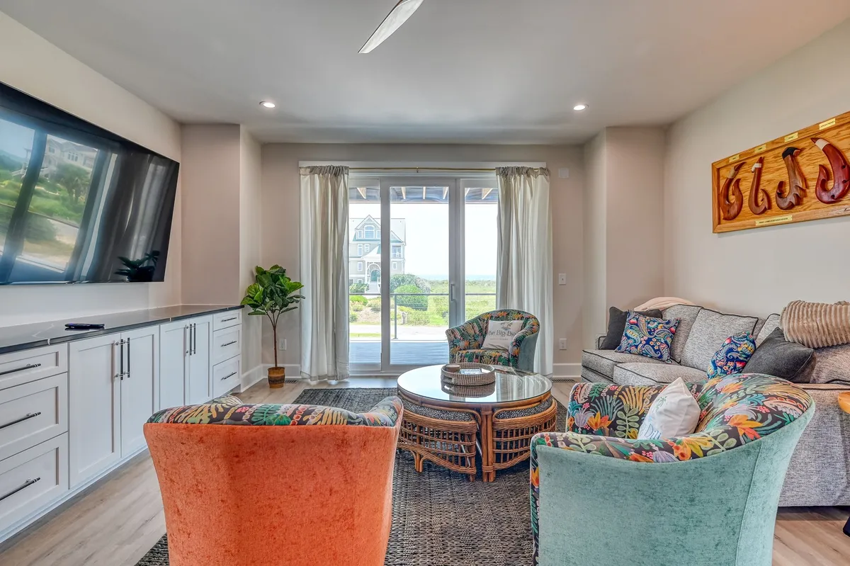 A vibrant, welcoming lounge with colorful armchairs arranged around a round coffee table, a large TV as the focal point, and sliding glass doors that bring in plenty of natural light. The space is designed for relaxing, socializing, or gathering for movie nights, and the overall decor feels lively yet comfortable.
